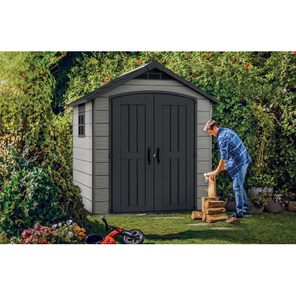 Na pierwszym planie mężczyzna rąbiący drewno, a za nim domek narzędziowy Premier 757 od firmy Keter