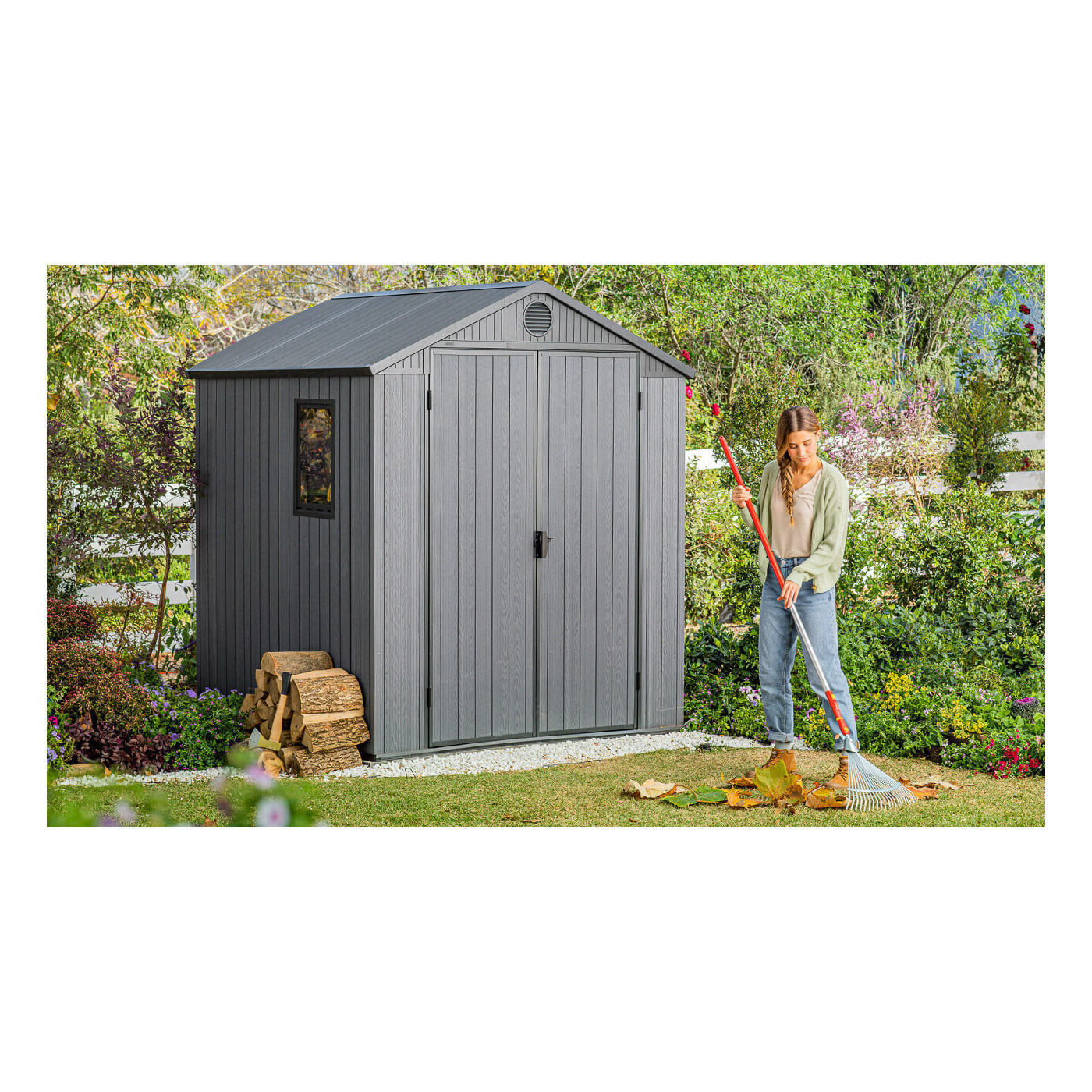 Na pierwszym planie kobieta grabiąca liście, a za nią stoi domek narzędziowy