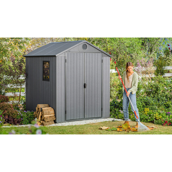 Na pierwszym planie kobieta grabiąca liście, a za nią stoi domek narzędziowy