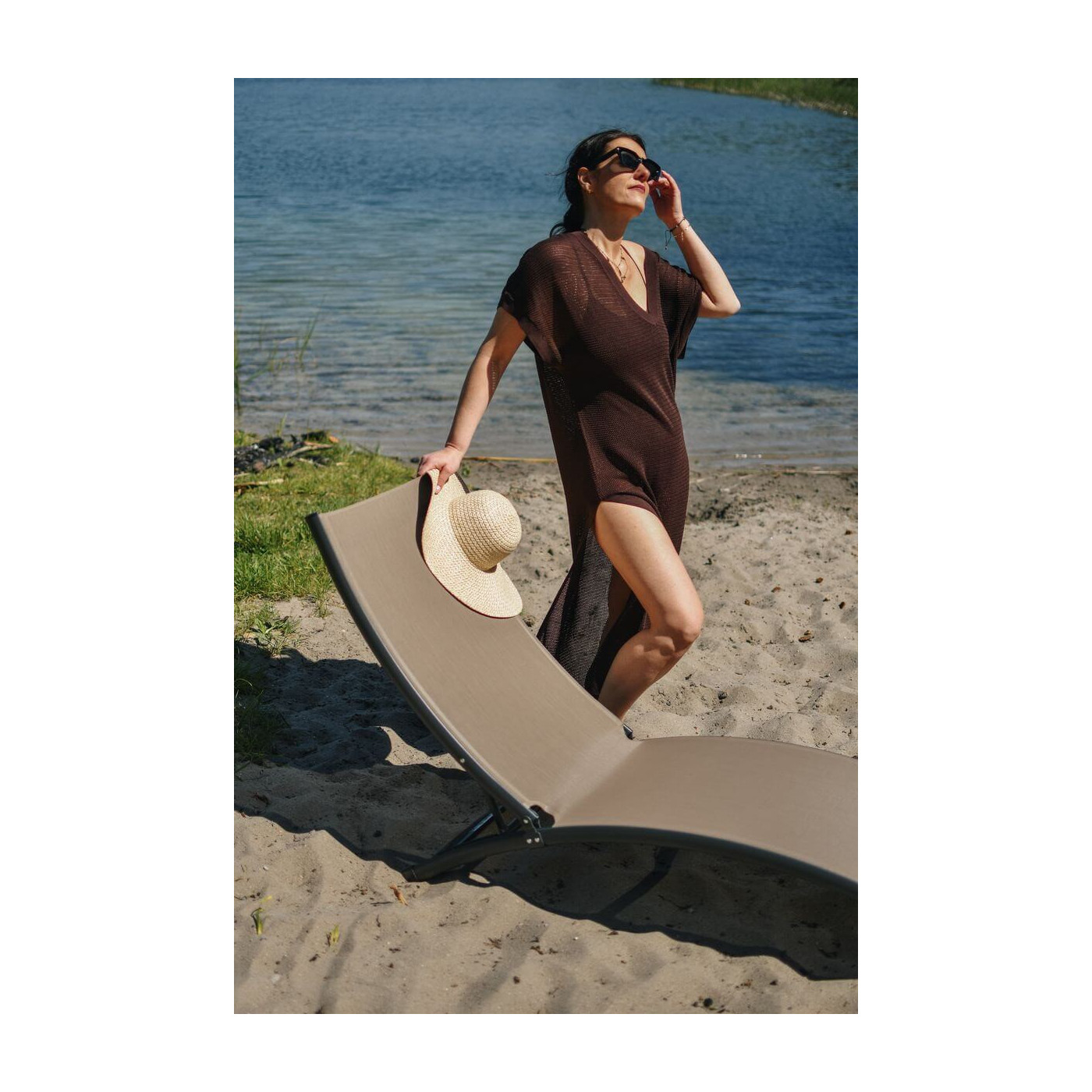 Kobieta oparta o leżak brązowy przy piaszczystym brzegu jeziora