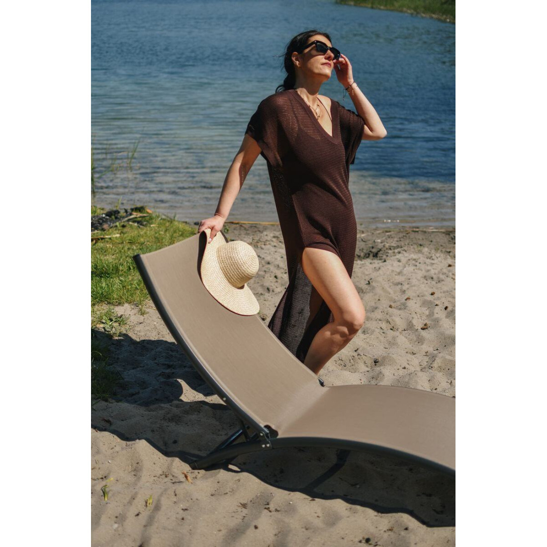 Kobieta oparta o leżak brązowy przy piaszczystym brzegu jeziora