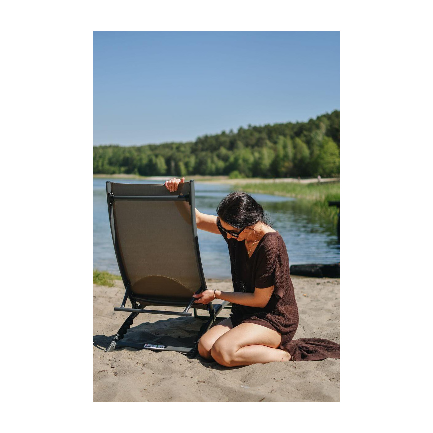 kobieta rozkładająca brązowy leżak Star na piasku przy jeziorze pośród drzew