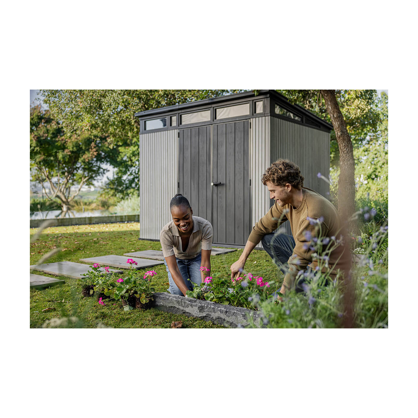 Kobieta i mężczyzna pielęgnują kwiaty w ogrodzie, a za nimi znajduje się antracytowa szopa na narzędzia