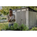 Na pierwszym planie kobieta dbająca o rośliny ogrodowe, a za nią stoi domek narzędziowy