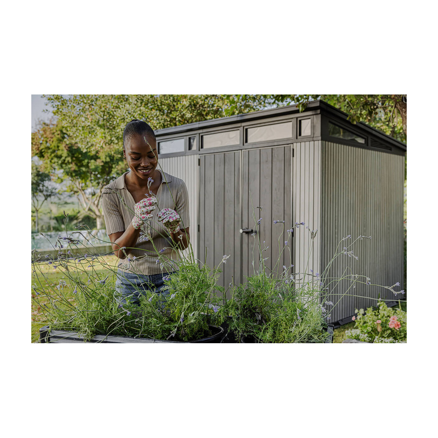 Na pierwszym planie kobieta dbająca o rośliny ogrodowe, a za nią stoi domek narzędziowy