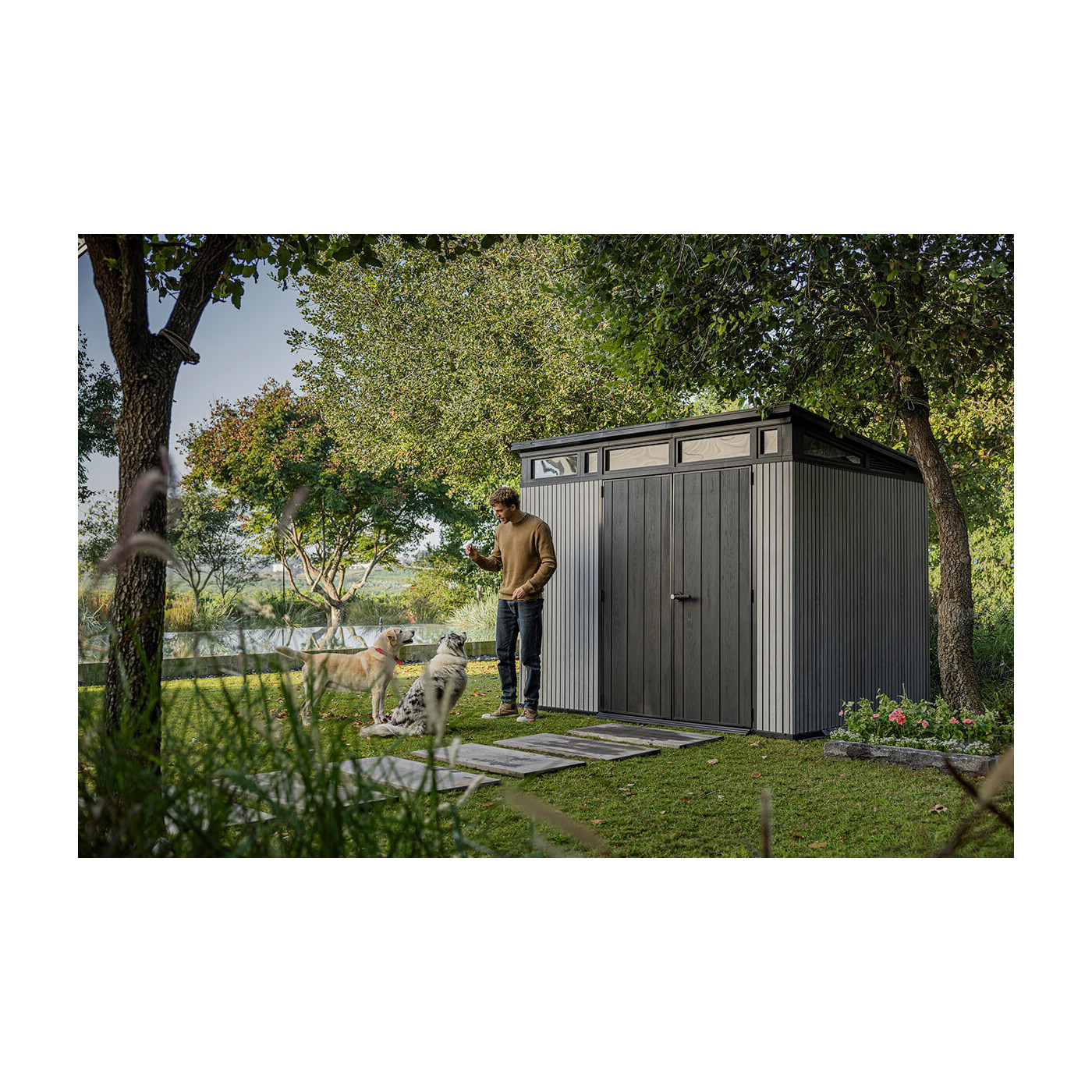 Aranżacja narzędziowni w ogrodzie, a obok niej mężczyzna bawiący się z psami