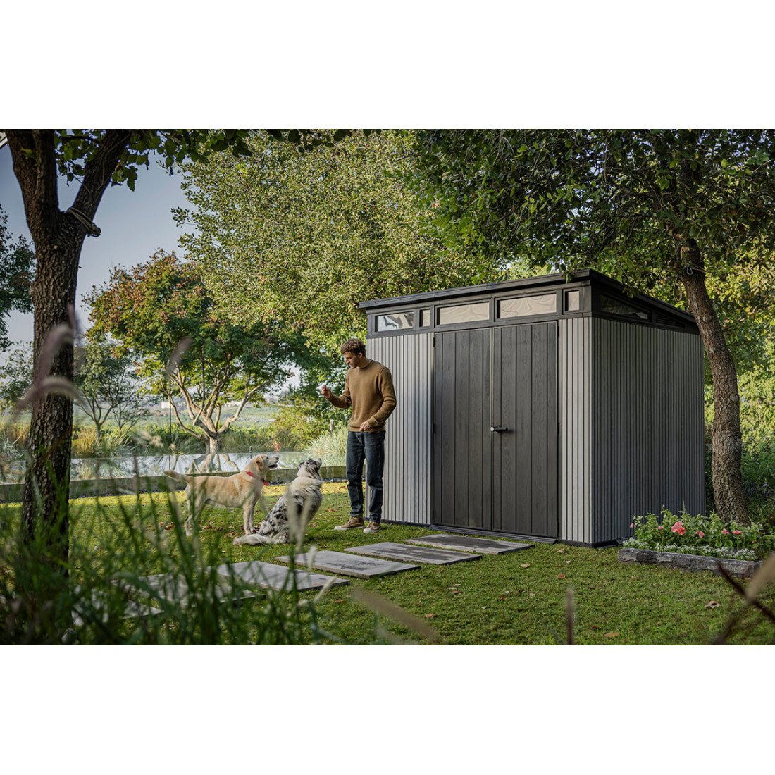 Aranżacja narzędziowni w ogrodzie, a obok niej mężczyzna bawiący się z psami