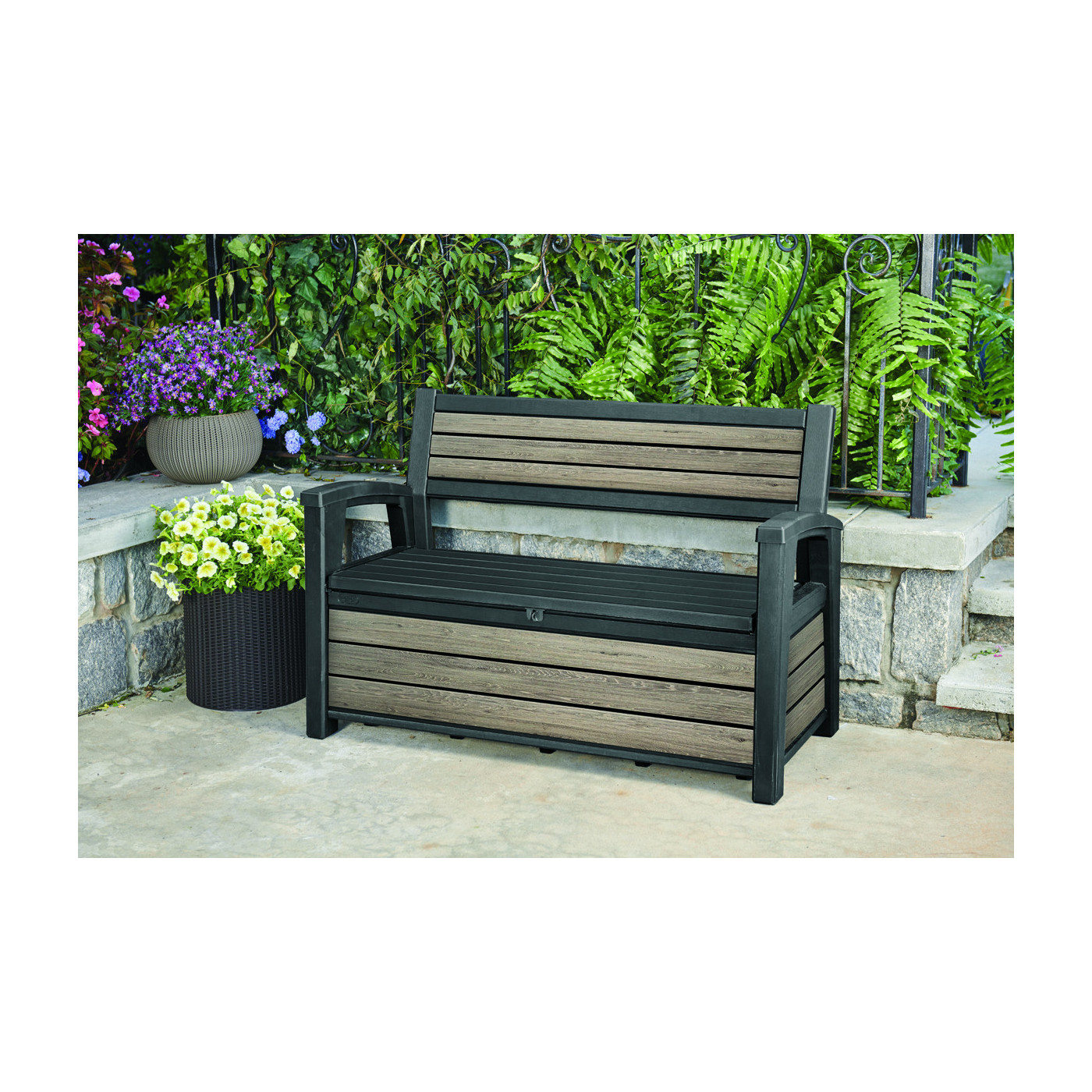 Ławka ogrodowa z pojemną skrzynią przy kamiennym murku obok dwóch donic z białymi i fioletowymi kwiatami 