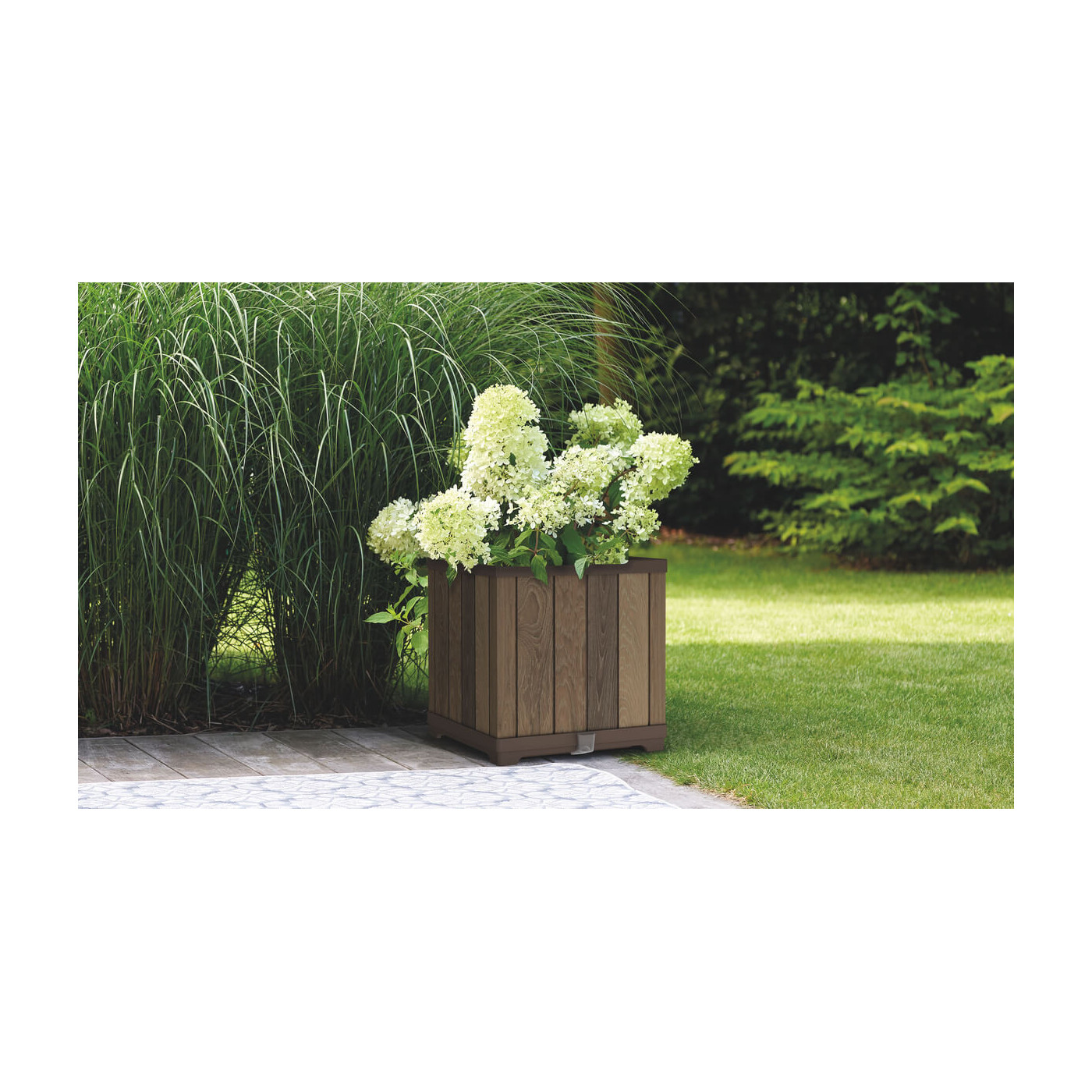 Duży, biały kwiat w kwadratowej, brązowej doniczce na zielonej trawie ,w tle drzewa iglaste i krzewy liściaste