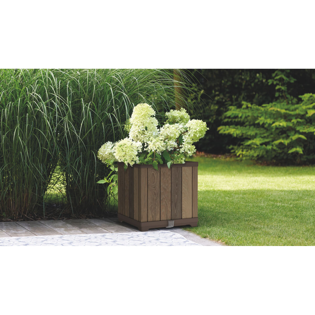 Duży, biały kwiat w kwadratowej, brązowej doniczce na zielonej trawie ,w tle drzewa iglaste i krzewy liściaste