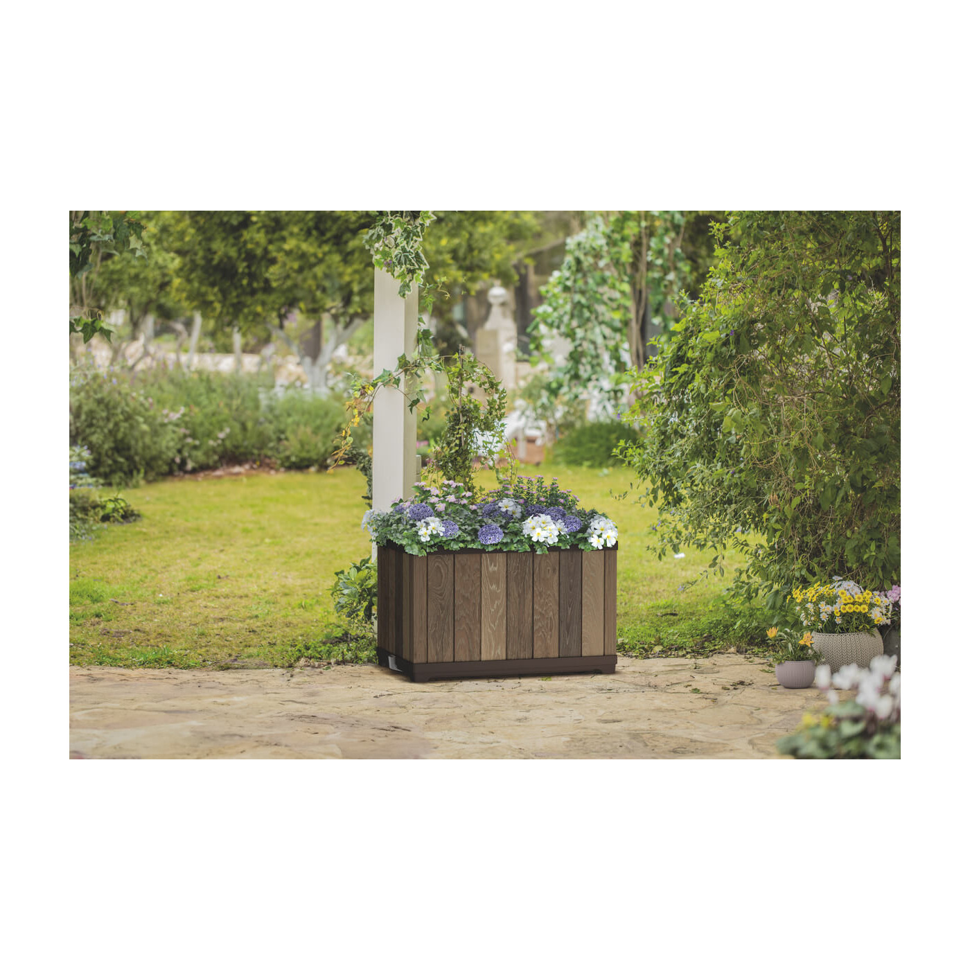 Elegancka donica w przydomowym ogrodzie otoczona kwiatami, krzewami i drzewami 